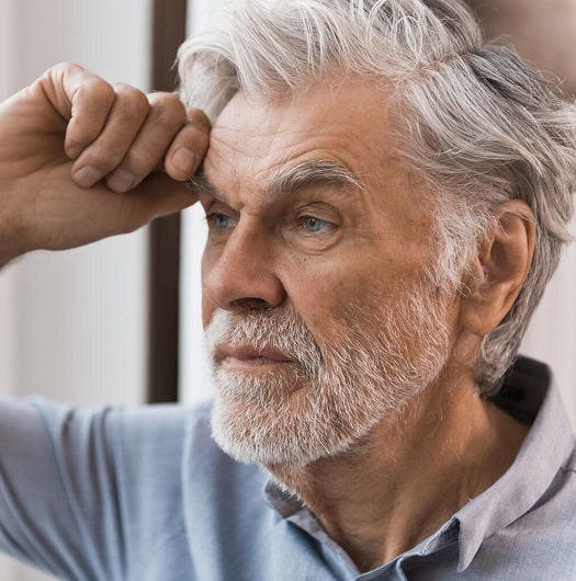 Une personne âgée aux cheveux gris ajuste un appareil auditif discret derrière son oreille droite. Elle se trouve dans un environnement lumineux, probablement une salle d’attente ou un intérieur calme.