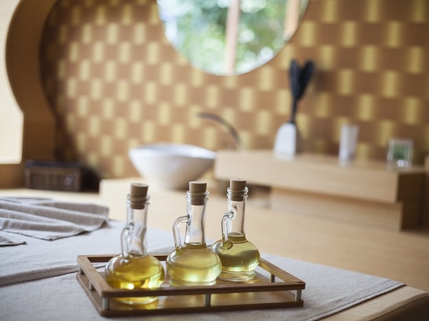 Trois flacons d’huile posés sur un plateau en bois, éclairés par une lumière naturelle, dans une pièce chaleureuse avec un lavabo moderne et une décoration aux tons dorés.