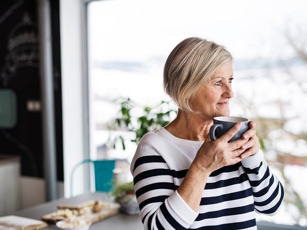 Une femme aux cheveux gris porte des aides auditives invisibles et boit une tasse de café dans une cuisine lumineuse.