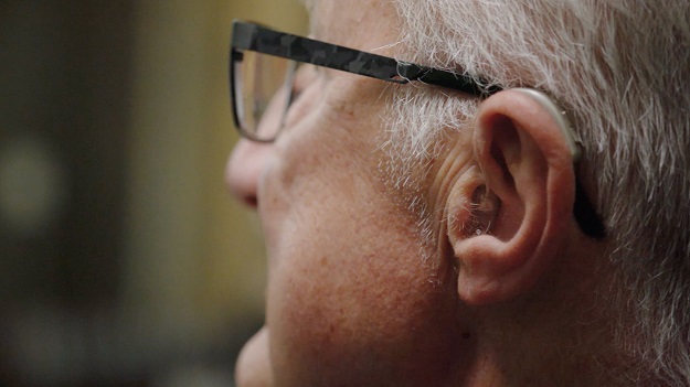 Vue rapprochée du profil d’un homme âgé portant un appareil auditif Audikafull et des lunettes.