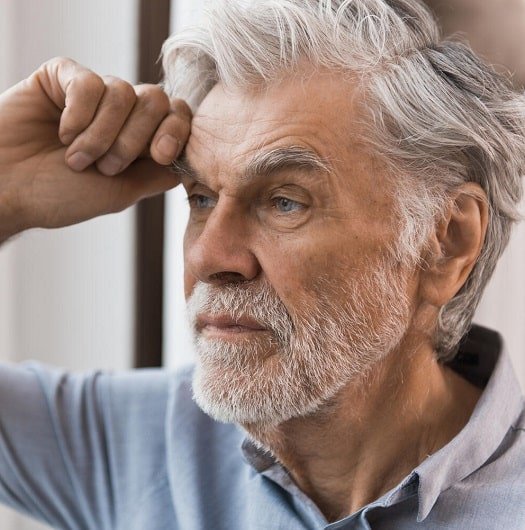 Une personne âgée aux cheveux gris ajuste un appareil auditif discret derrière son oreille droite. Elle se trouve dans un environnement lumineux, probablement une salle d’attente ou un intérieur calme.