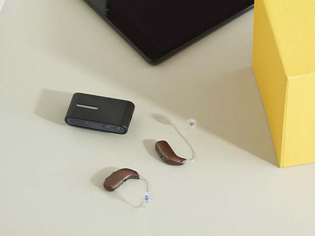 Deux aides auditives marron à contour d’oreille (type Oticon) reposent sur une surface beige, accompagnées d’un accessoire noir connecté; en arrière-plan, une tablette et une boîte jaune.