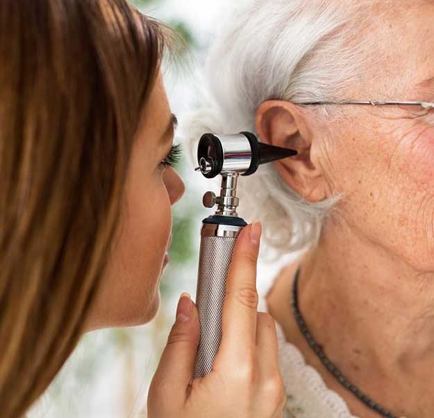 Une personne âgée aux cheveux blancs reçoit un examen auditif avec un otoscope tenu par un audiologiste dans un environnement lumineux, probablement un centre auditif ou une clinique spécialisée.