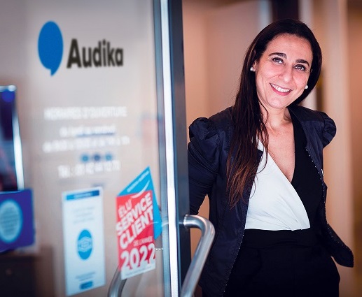 Femme entrant dans un centre auditif Audika avec des informations sur les heures d’ouverture visibles sur la porte.