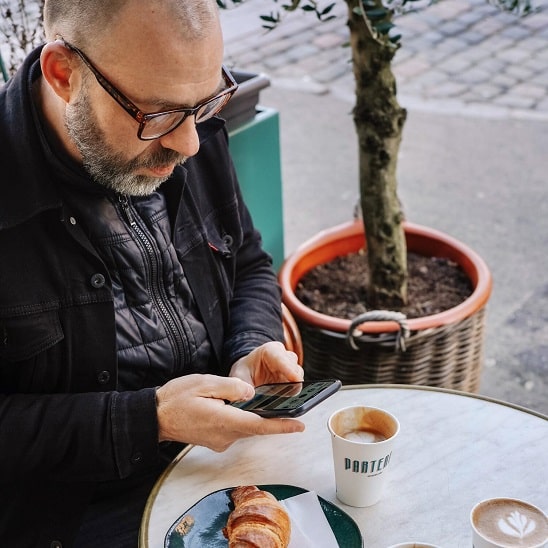 Un client utilisant un appareil auditif Audika laisse un avis sur son téléphone en terrasse avec un café et un croissant.