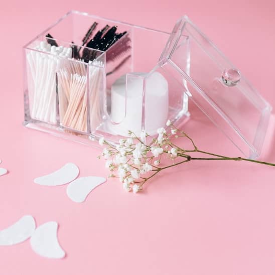 Bâtonnets ouatés et accessoires de beauté dans un organiseur transparent sur fond rose avec fleurs décoratives.