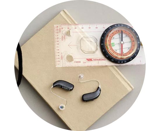Appareils auditifs Audikafull noirs posés sur un carnet beige à côté d'une boussole et d'une règle transparente.
