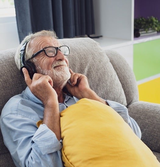 Un homme âgé assis sur un canapé écoute avec un casque audio, tenant un coussin jaune.