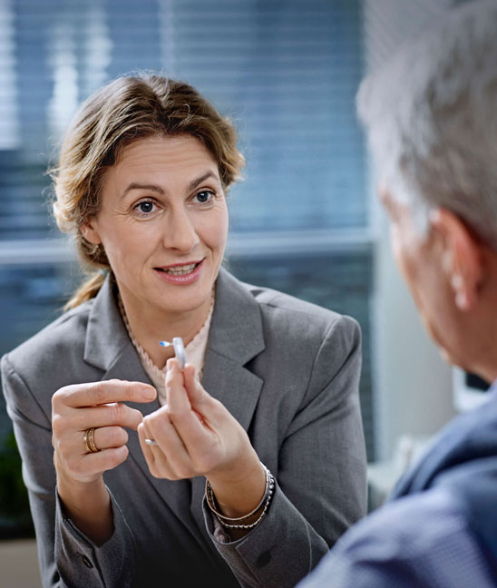 Une audioprothésiste montre une aide auditive personnalisée à un patient lors d’une consultation.
