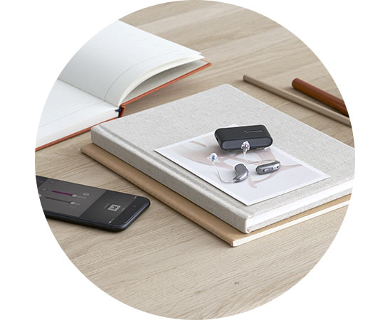 Aides auditives modernes avec leur boîtier de charge, disposées sur un carnet sur un bureau en bois.