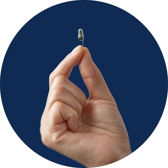 Une main tient une aide auditive invisible de type intra-auriculaire devant un fond bleu