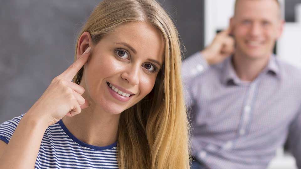 Una donna indica il proprio orecchio con il dito, segnalando un possibile problema uditivo; sullo sfondo, un uomo fa un gesto simile.