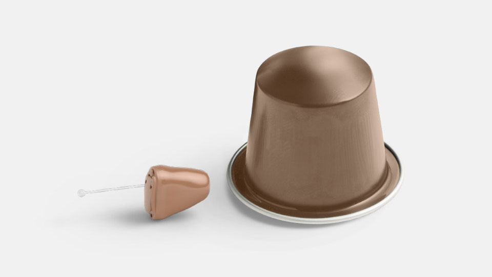 Un piccolo apparecchio acustico color carne è posizionato accanto a una capsula da caffè marrone su uno sfondo bianco, evidenziando le sue dimensioni discrete.