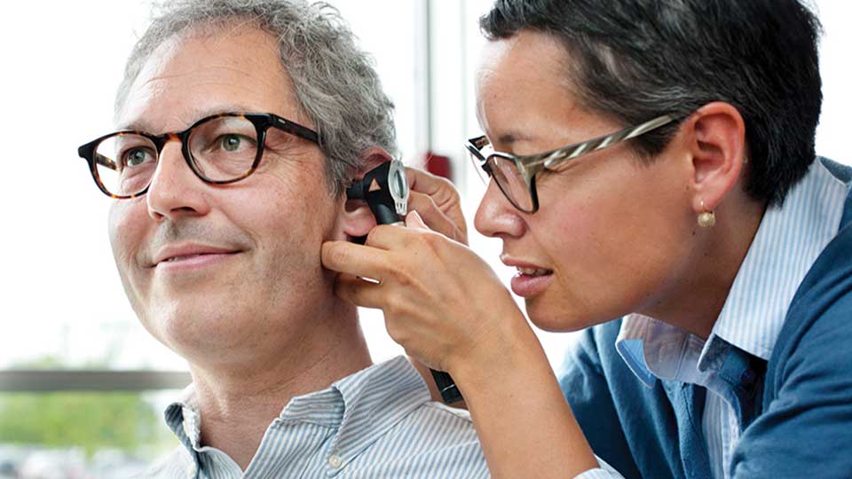 Un audioprotesista esamina l'orecchio di un uomo con un otoscopio, all'interno di un centro acustico Audika. Lo sfondo mostra una finestra con luce naturale.