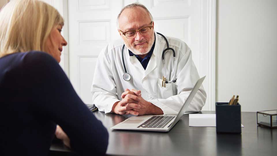 Un medico con camice bianco e stetoscopio al collo interagisce con una paziente in una stanza luminosa. Sul tavolo ci sono un laptop, un portapenne e documenti.