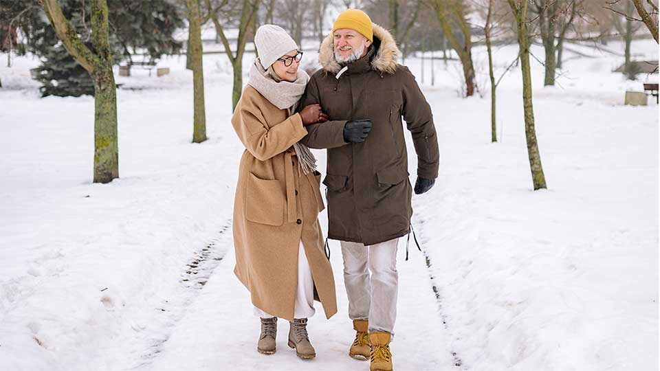 Una coppia cammina abbracciata su un sentiero innevato in un parco, indossando cappotti invernali; intorno, alberi spogli e un paesaggio tranquillo creano un’atmosfera serena.