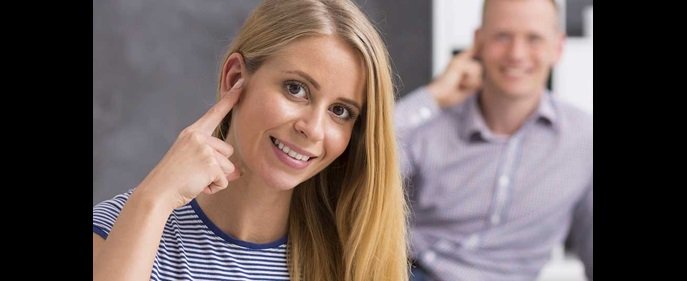 Una donna indica il proprio orecchio con il dito, segnalando un possibile problema uditivo; sullo sfondo, un uomo fa un gesto simile.