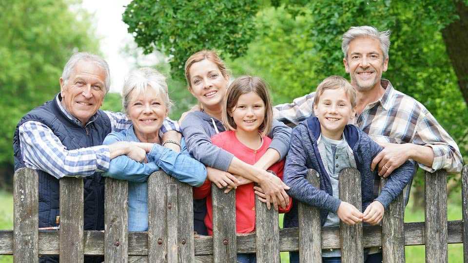 Un gruppo familiare, composto da adulti e bambini, si appoggia rilassatamente a una staccionata in legno. Sullo sfondo, un ambiente naturale con prati verdi e alberi.