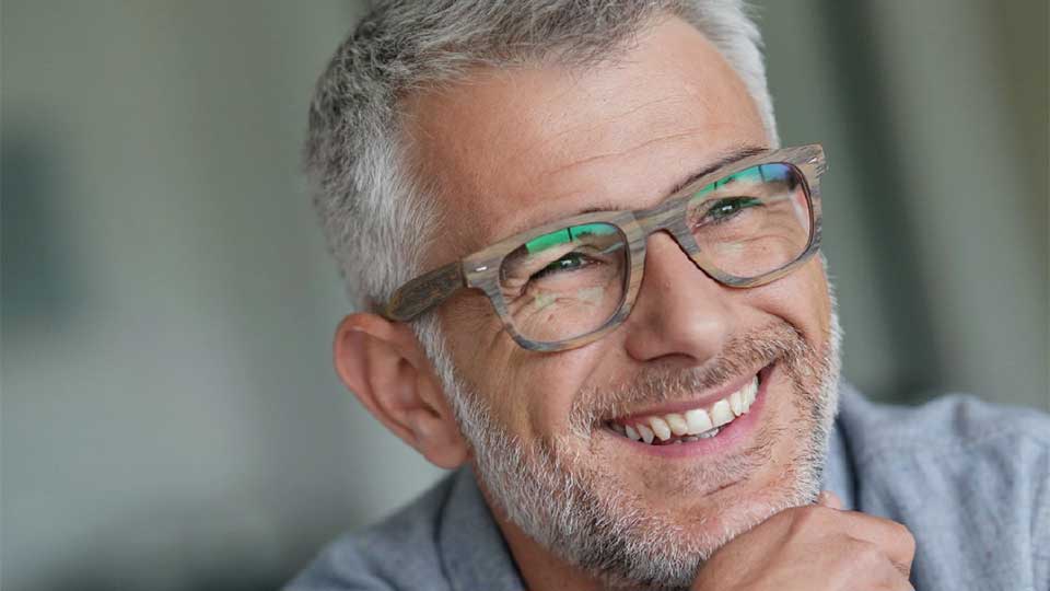 Un uomo con capelli grigi indossa un apparecchio acustico dietro l’orecchio sinistro. Sullo sfondo, sfocato, si intravede un ambiente interno dai toni neutri.