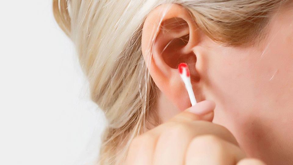 Un orecchio femminile viene pulito con un bastoncino di cotone, visibile del sangue sulla punta. Lo sfondo è chiaro, con focus sulla zona auricolare.
