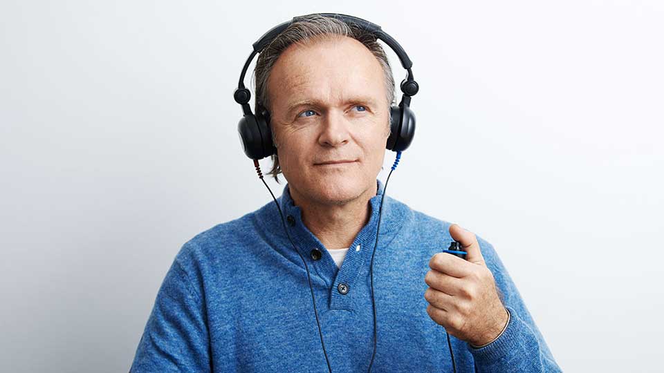 Un uomo con una maglia blu indossa delle cuffie nere e tiene un pulsante durante un test dell’udito in un ambiente clinico, presso un centro acustico Audika.