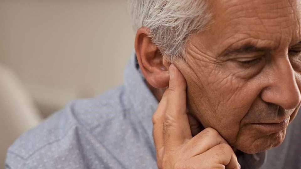 Un uomo anziano con capelli grigi, indossando una camicia azzurra, tocca l’orecchio sinistro. Il contesto suggerisce un ambiente interno, forse un centro acustico. 