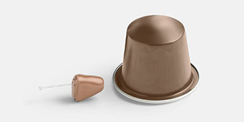Un piccolo apparecchio acustico color carne è posizionato accanto a una capsula di caffè marrone su uno sfondo bianco, evidenziando le dimensioni discrete dell'apparecchio.