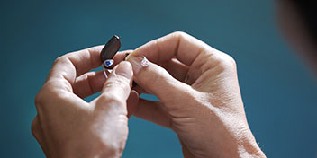 Mani umane tengono e regolano un piccolo apparecchio acustico intrauricolare in un ambiente con sfondo sfocato blu, suggerendo un contesto clinico o una consulenza uditiva professionale.
