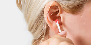 Una donna bionda utilizza un cotton fioc per pulire l'interno dell'orecchio in un contesto neutro con sfondo bianco, richiamando l'importanza della cura dell'udito.