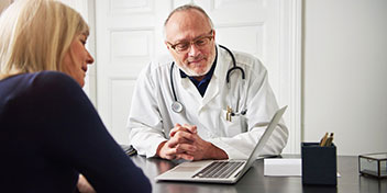 Un medico con camice bianco e stetoscopio è seduto di fronte a una donna in una consulenza presso una clinica, con un laptop e una scatola sul tavolo.