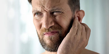 Un uomo tocca il proprio orecchio con la mano, suggerendo un possibile problema uditivo. Lo sfondo è sfocato, con tonalità chiare che trasmettono un ambiente neutro o clinico.