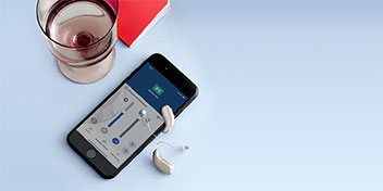 Un apparecchio acustico moderno accanto a uno smartphone che mostra l’app di controllo del dispositivo (schermata con cursori e opzioni), un bicchiere e un libro su sfondo chiaro.