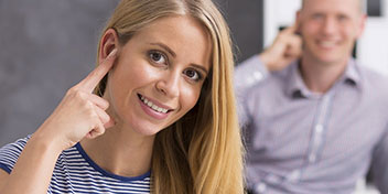 Una donna indica il suo orecchio con un dito, suggerendo un contesto legato a problemi uditivi. Sullo sfondo, un uomo sta toccando l’orecchio. Ambiente tipico di un centro acustico.