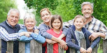 Un gruppo familiare, composto da adulti e bambini, posa appoggiato a una recinzione di legno in un ambiente naturale con alberi e prati verdi sullo sfondo.