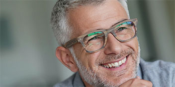 Un uomo con capelli grigi indossa un maglione grigio in un contesto domestico sfocato. L’immagine si collega alla cura dell’udito.