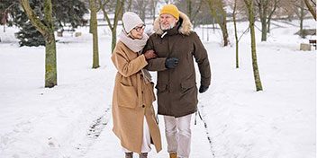 Una coppia matura cammina sorridendo su un sentiero innevato, abbracciata calorosamente. Indossano cappotti invernali e cappelli; lo sfondo mostra alberi e un parco innevato.