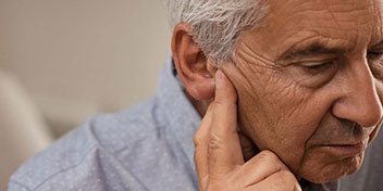 Un uomo anziano, con capelli grigi e camicia azzurra, mostra un orecchio nudo senza apparecchio acustico visibile. Lo sfondo è sfocato, suggerendo un ambiente interno.