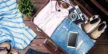 Una valigia aperta contiene una camicia rosa, jeans, una fotocamera vintage, scarpe e uno smartphone; intorno ci sono vestiti sparsi e un vaso di pianta su un pavimento in legno.