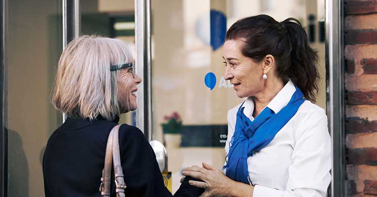 Una donna con capelli grigi e borsa beige interagisce con una professionista in camicia bianca e foulard blu davanti a un centro acustico Audika; porte di vetro mostrano l’interno accogliente.