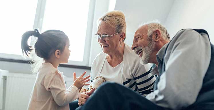 Una bambina parla con due anziani seduti su una sedia in una stanza luminosa con ampie finestre. L’atmosfera è intima e familiare. 
