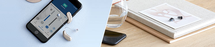 Due apparecchi acustici Audika beige accanto a uno smartphone con app di controllo aperta, su superficie chiara. A destra, un tavolo con libri, bicchiere e apparecchi acustici neri.