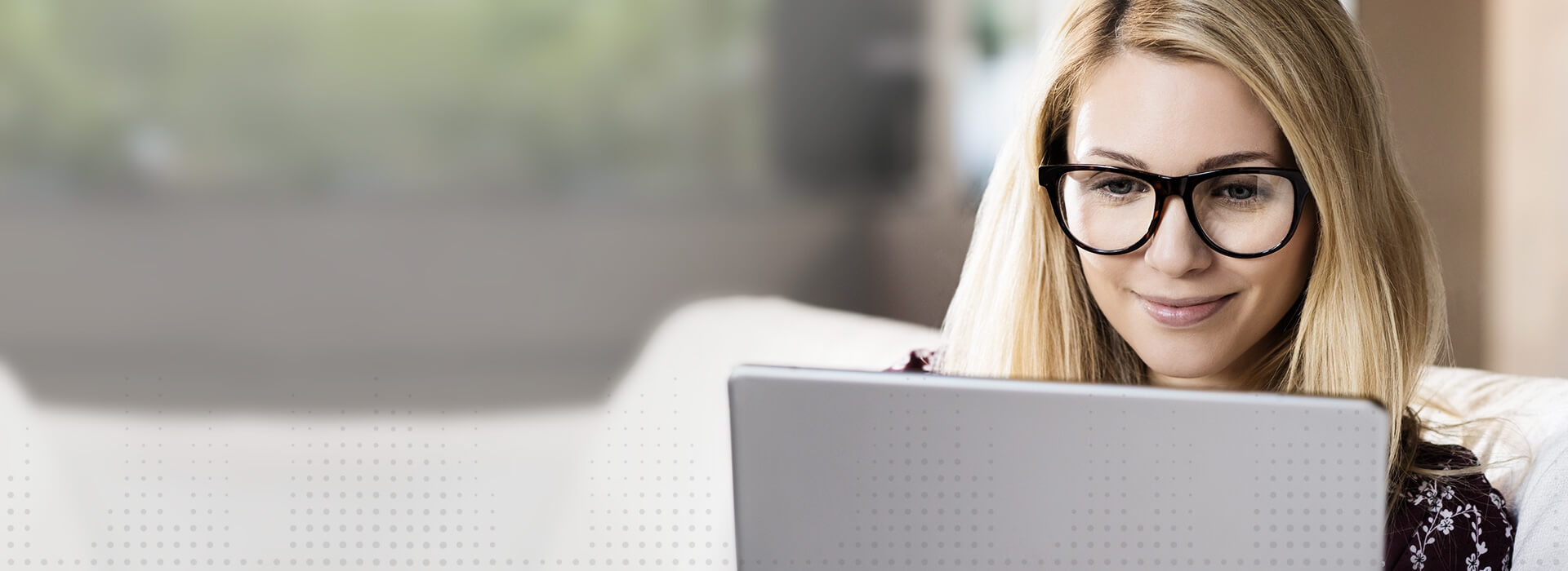 Una donna con lunghi capelli biondi legge o lavora su un dispositivo elettronico in un ambiente domestico luminoso con sfondo sfocato e accogliente.