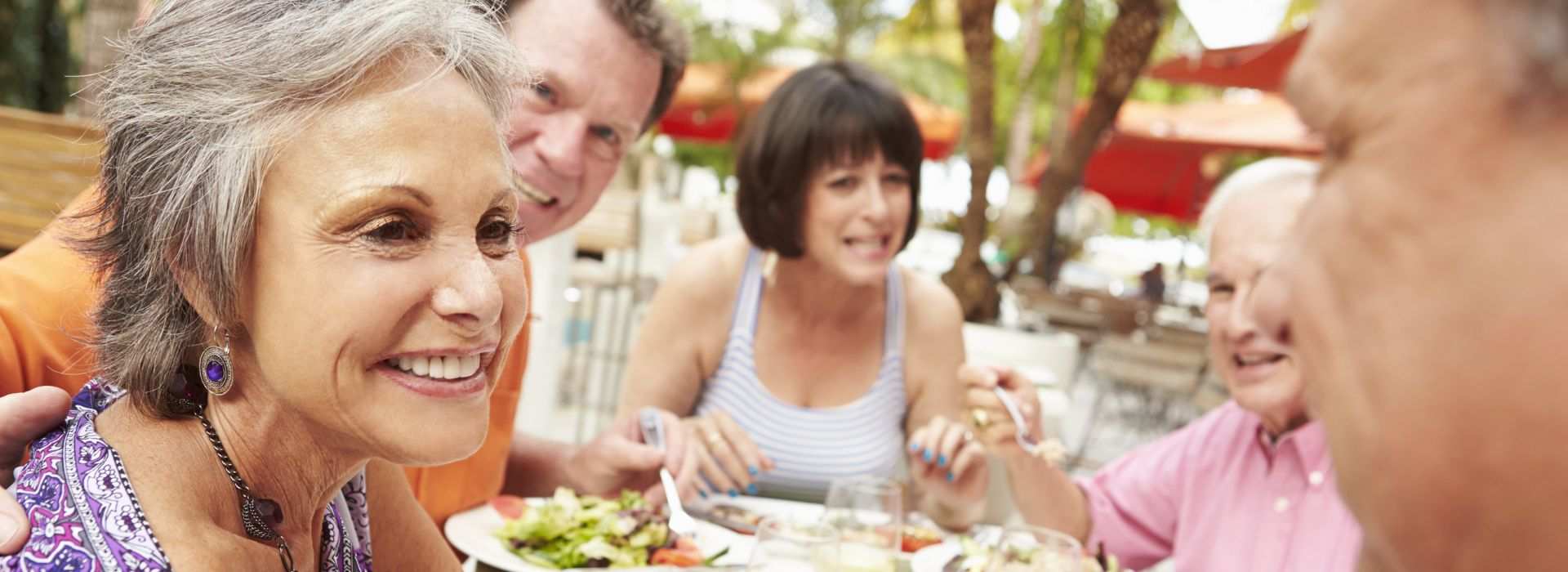 Anziana con capelli grigi che indossa un apparecchio acustico retroauricolare mentre partecipa a un pranzo all’aperto.