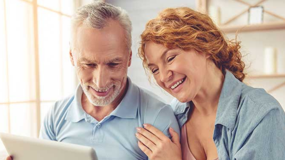 Un uomo anziano con una polo azzurra tiene un tablet, mentre una donna dai capelli ricci rossi gli appoggia una mano sulla spalla. Sfondo: interno luminoso con scaffali eleganti.