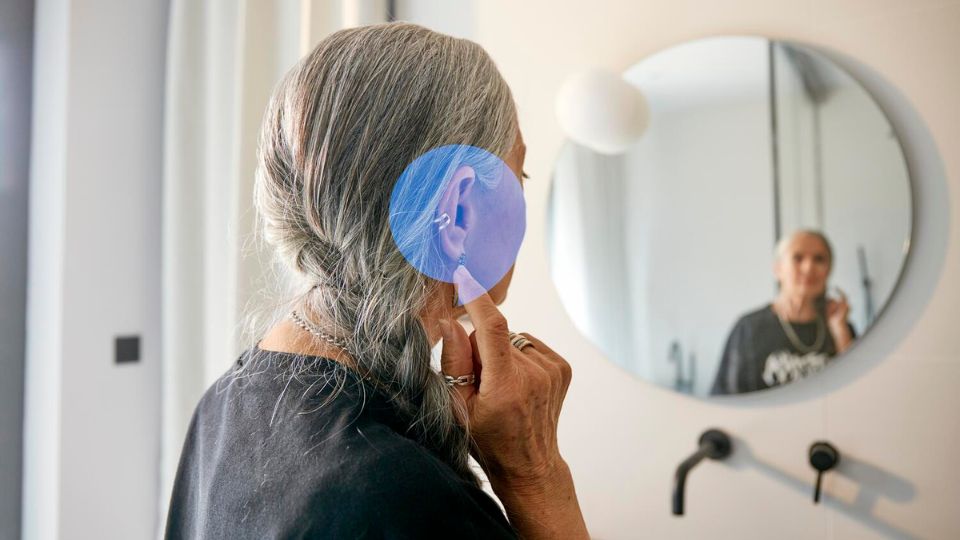 Donna anziana con capelli grigi che indossa un apparecchio acustico, riflessa in uno specchio del bagno.