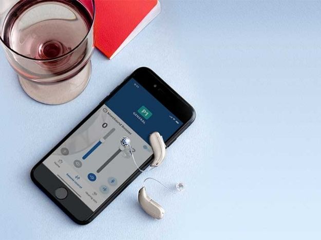 Due apparecchi acustici sono posizionati accanto a uno smartphone che mostra un'app di controllo per regolare impostazioni uditive; un bicchiere e un taccuino rosso sullo sfondo.