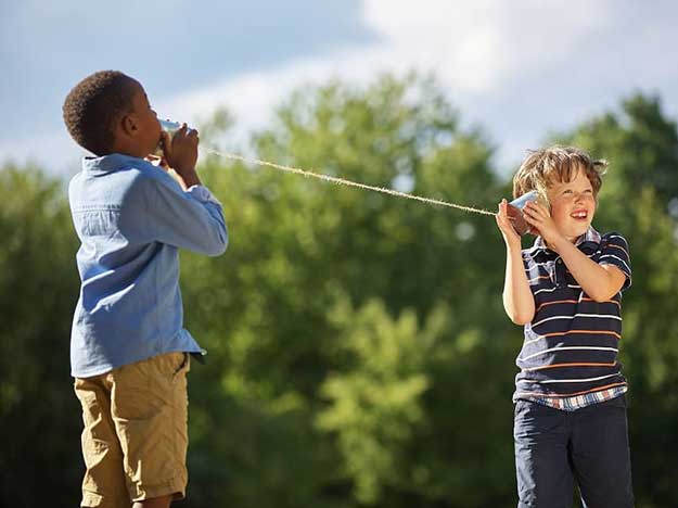 Due bambini giocano con un telefono di latta collegato da una corda, in un parco verdeggiante con alberi sullo sfondo sotto un cielo parzialmente nuvoloso.