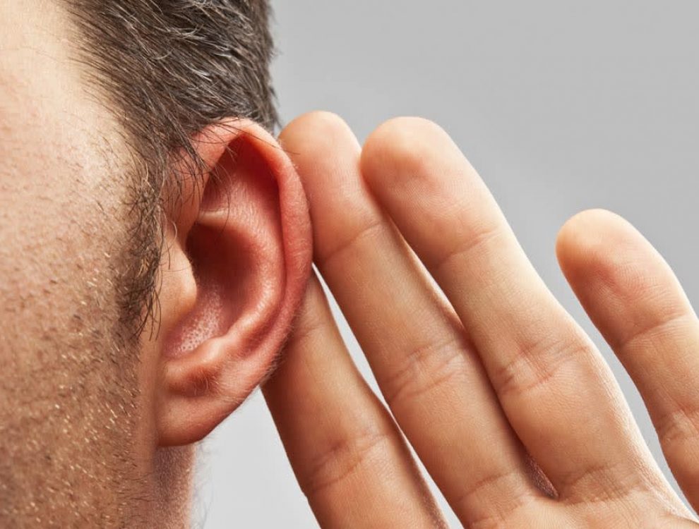 Un orecchio maschile in primo piano viene avvicinato a una mano a forma di conca, suggerendo difficoltà uditive. Sfondo grigio neutro. Contesto ideale per promuovere test dell’udito.