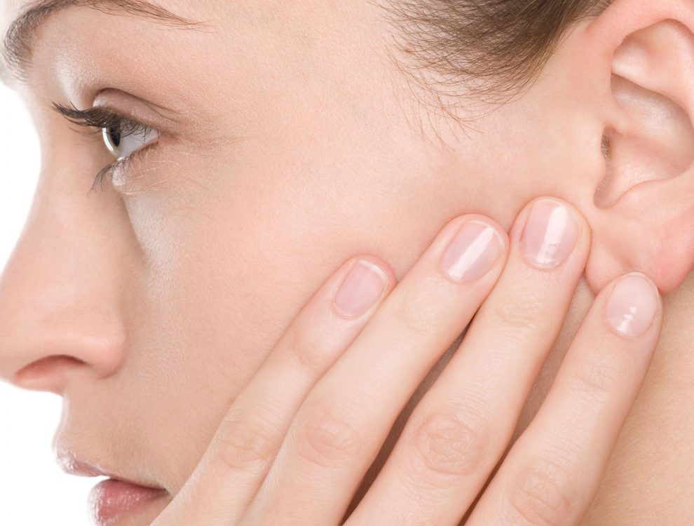 Orecchio umano tenuto delicatamente con due dita, sfondo sfumato color carne. Il contesto suggerisce un focus sull’udito o la cura dell’orecchio, come in un centro acustico.