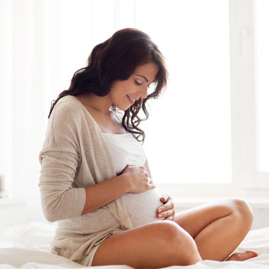 Una donna incinta, seduta con le gambe incrociate su un letto, tiene le mani sul pancione. Indossa abiti chiari e si trova in una stanza luminosa con luce naturale.